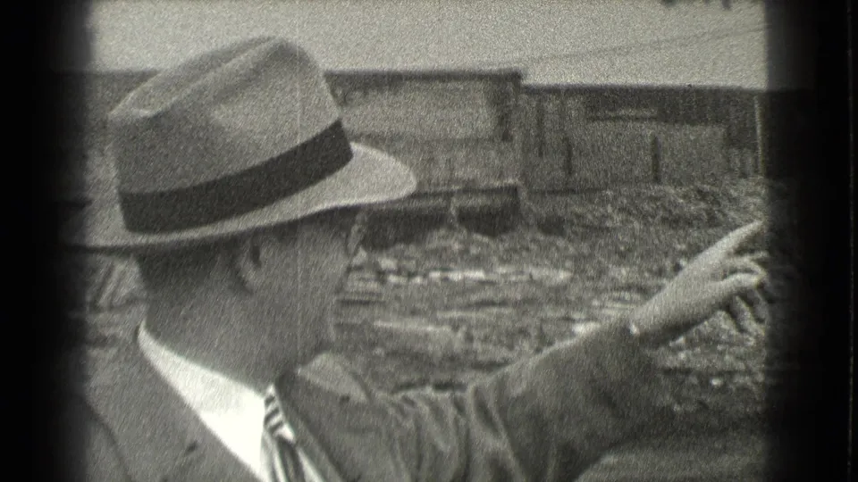 The 1939 Cleveland footage shows the man turning and pointing across a debris-strewn construction site with a long unfinished structure behind him.