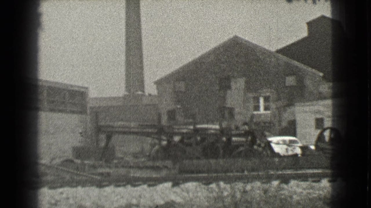 Industrial buildings beside train tracks with a smokestack and parked cars.