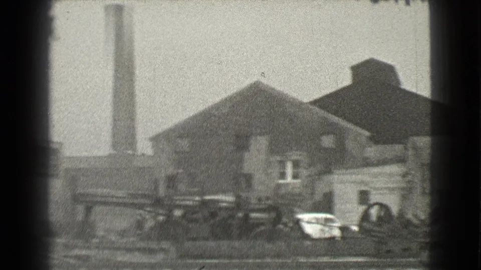 1939 Cleveland home-movie frame showing a brick industrial building, tall smokestack, and cluttered yard foreground.