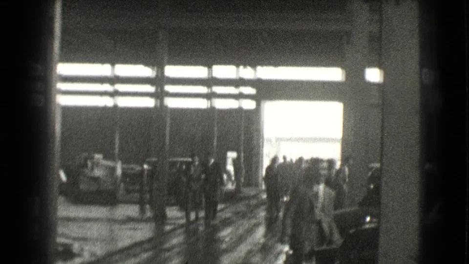 1939 Cleveland interior frame showing people gathered in a large dim hall with vehicles and bright exterior light beyond the doorway.