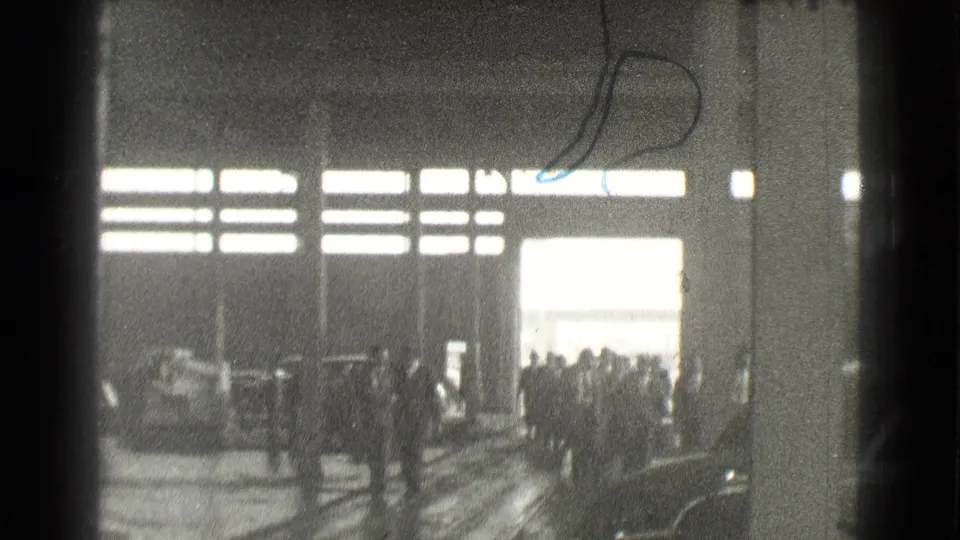 Black-and-white 8mm frame of a Cleveland hall interior where a small crowd stands near the light from an open doorway.
