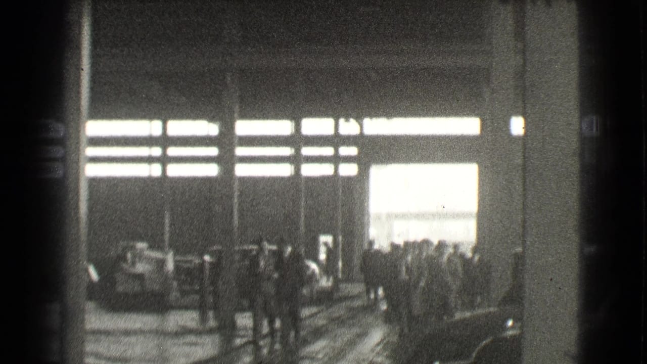 Dim factory interior with workers gathered near bright high windows.