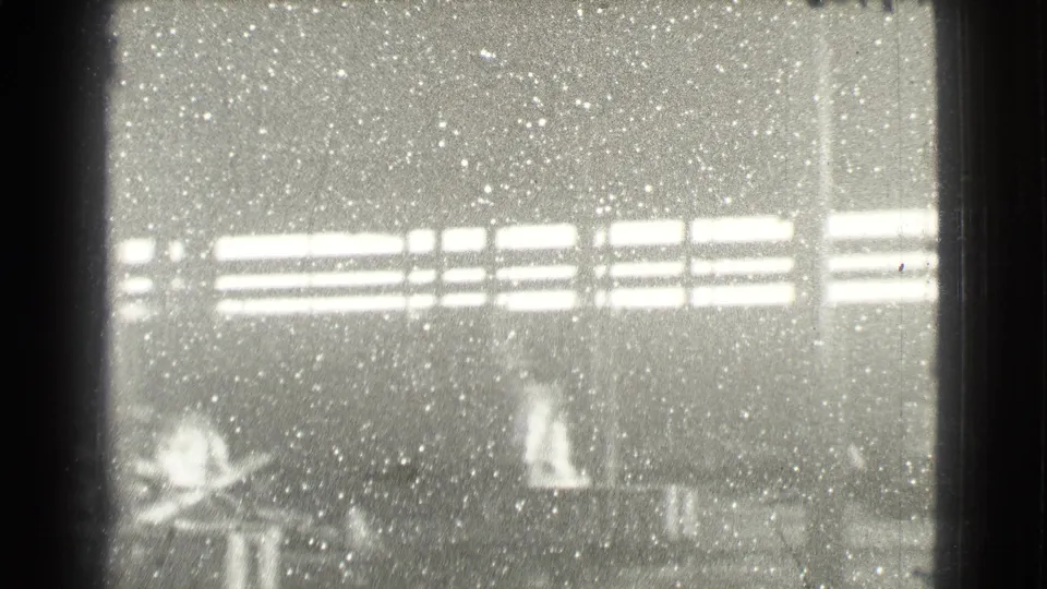 Overexposed 8mm frame of a Cleveland factory interior, with window glare, grain and faint figures or work surfaces below.