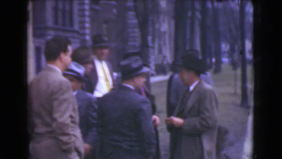 1939 Cleveland home-movie frame showing men in suits and fedoras gathered in conversation beside stone building fronts.