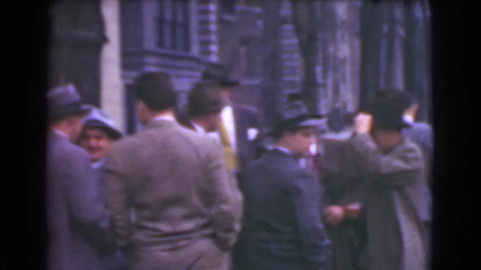 1939 Cleveland street frame with suited men and hat-wearing adults clustered in conversation beside urban building fronts.