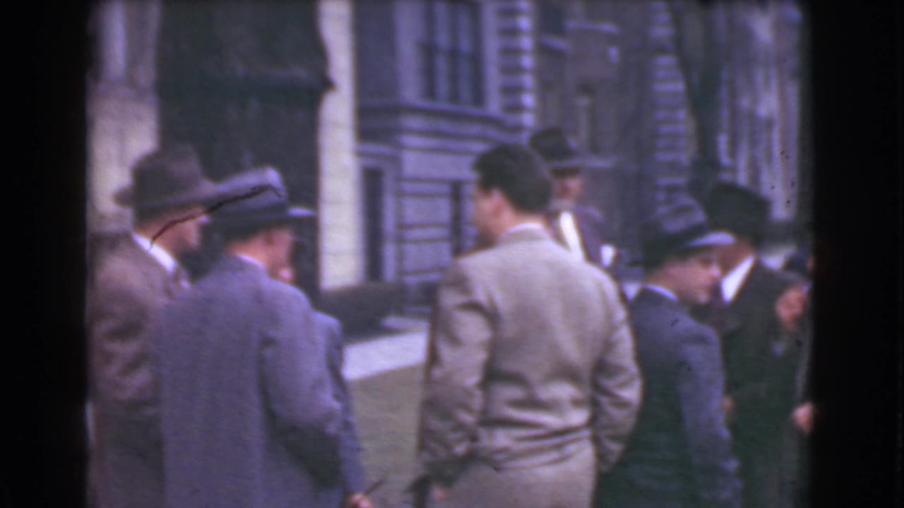 Men in suits and hats stand in a sidewalk group outside a stone building.