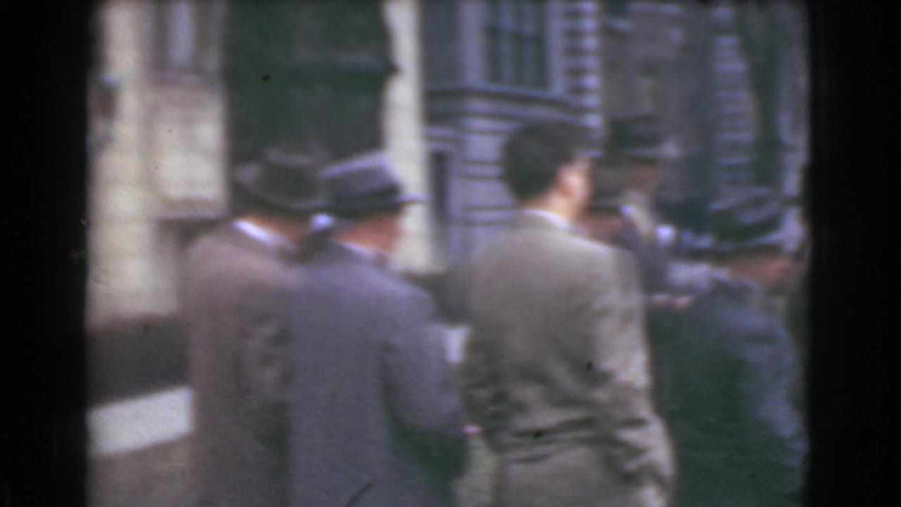 Men in suits and hats seen from behind on a city sidewalk near a stone building.