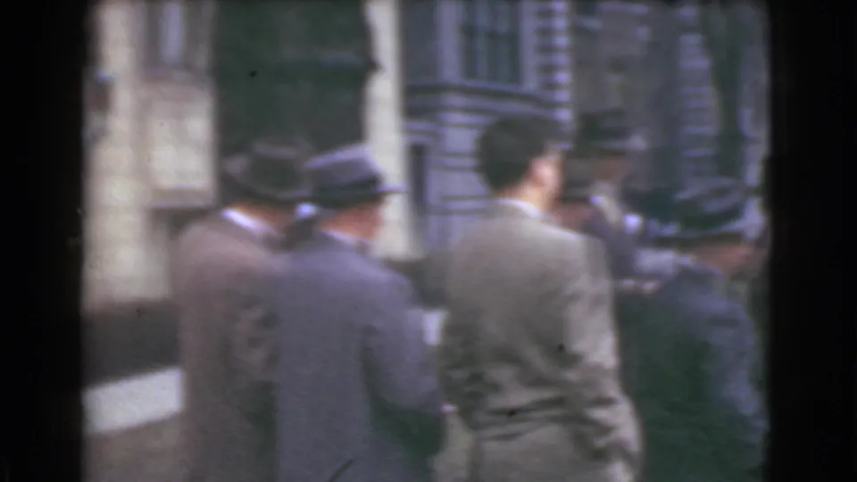 Cleveland 1939 home-movie frame showing adult men in suits and brimmed hats gathered on a city sidewalk beside stone architecture.