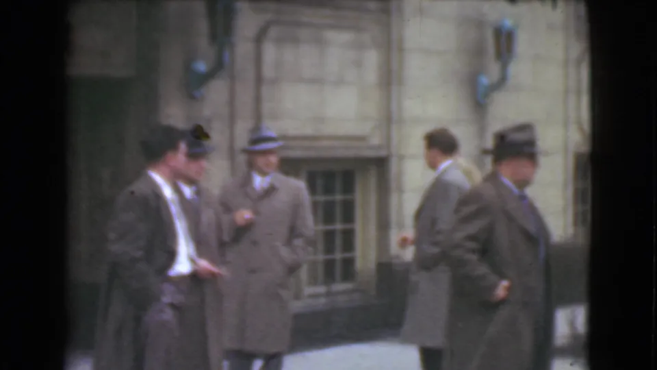 A short Cleveland 1939 sidewalk view shows suited men in hats, with one man walking past others near a stone facade.