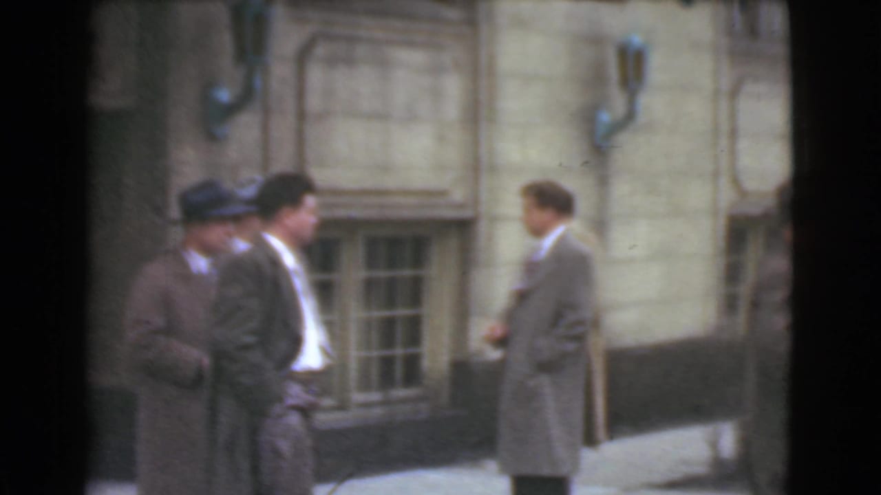 Men in suits and hats stand outside a stone building with lower windows.