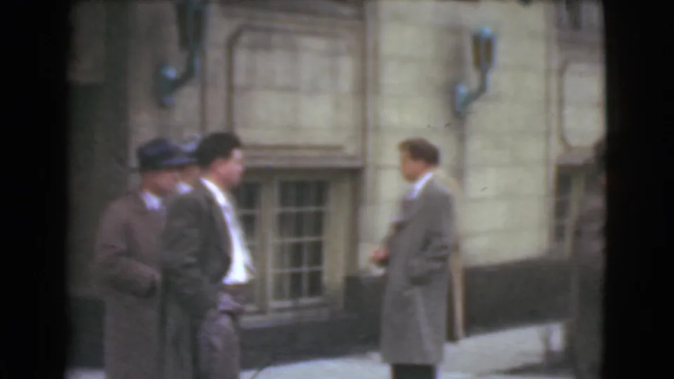 In Cleveland archival home-movie footage from 1939, adult men in suits, overcoats, and hats stand near a stone facade.