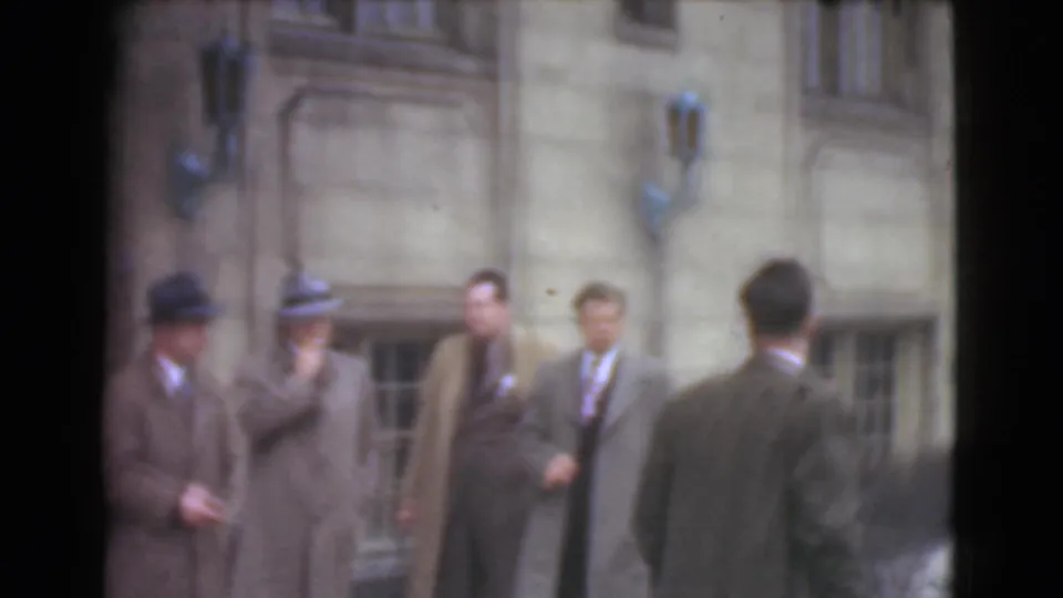 A suited man seen from behind faces other adult men near the stone building exterior in Cleveland home-movie footage.