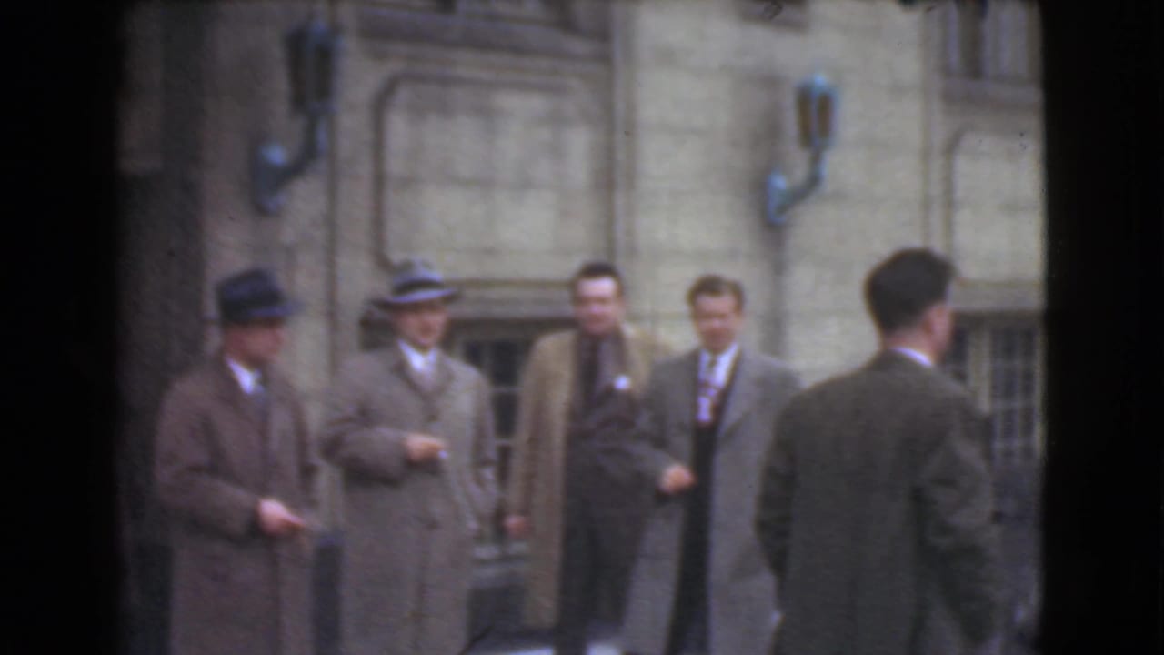 Men in suits and hats stand outside a masonry building, with one man seen from behind.