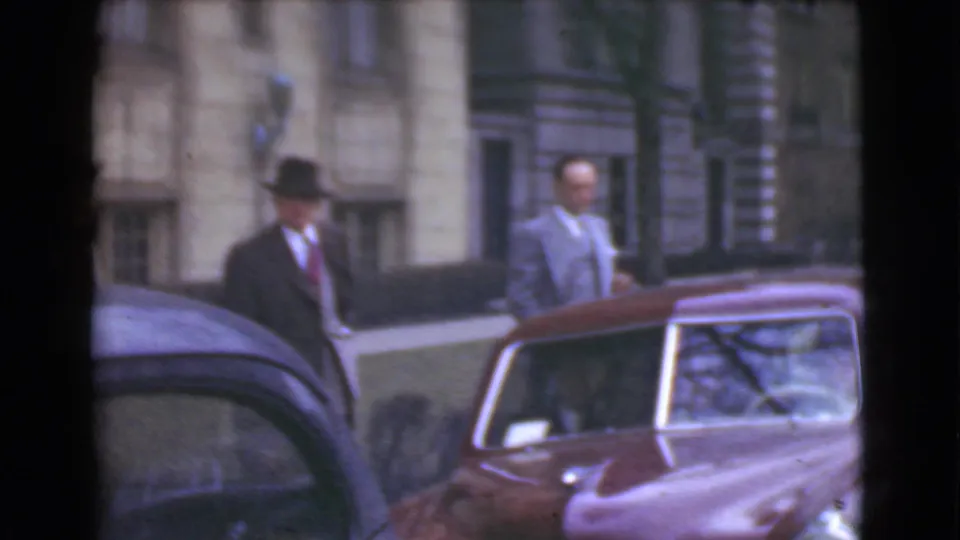 Men in period suits stand beside parked automobiles outside a Cleveland street building, including a maroon car in the foreground.