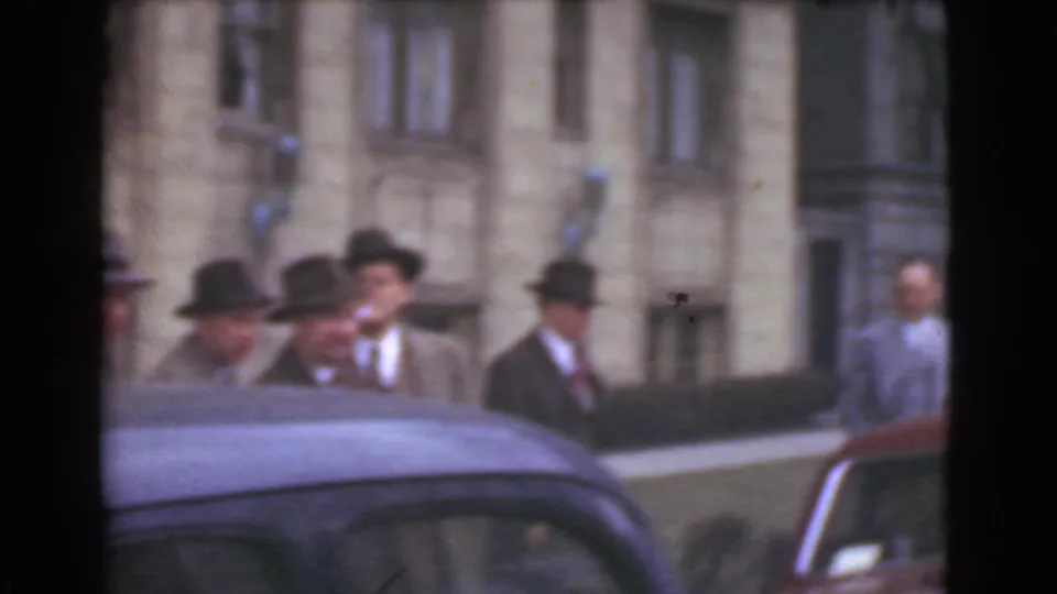 The frame shows suited men in dark hats partly obscured by a parked car, with a city building facade behind them.