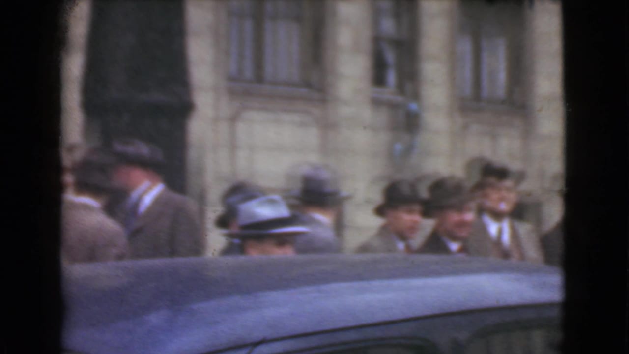 Men in suits and hats stand near a stone building with a dark car roof in the foreground.