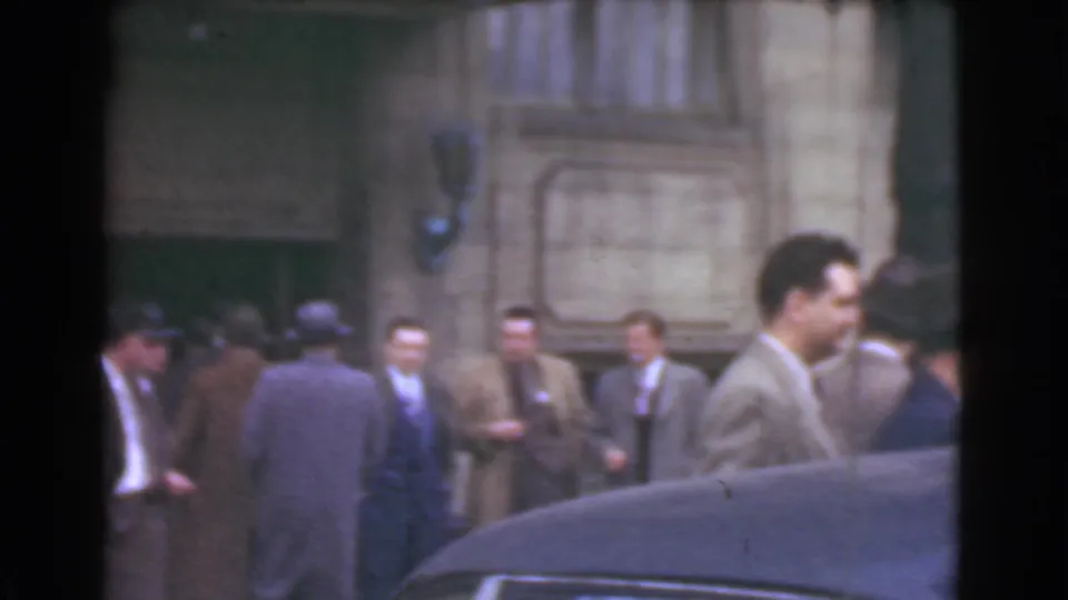 Suited men in hats gather and move near a stone building entrance, with a dark car roof cutting across the lower frame.