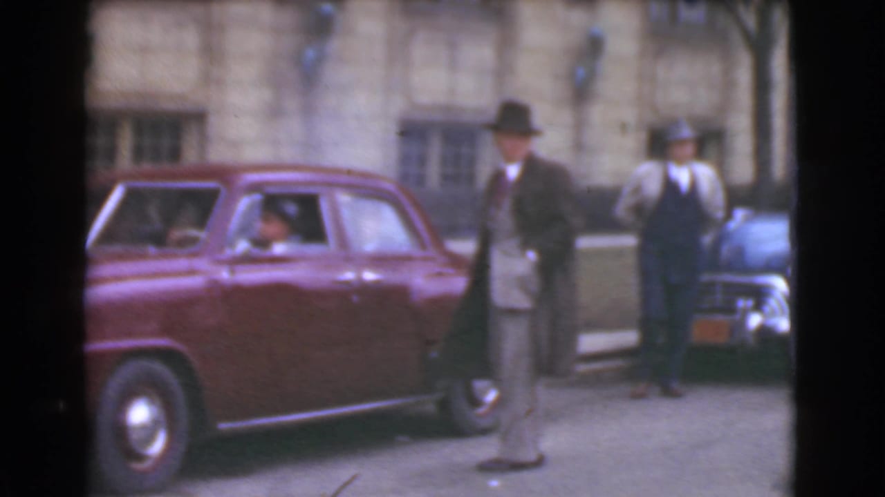 Men in fedoras stand beside a maroon sedan on a city street.