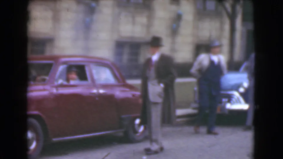 The maroon sedan moves through the Cleveland street scene as two men in hats stand near a blue car and stone facade.