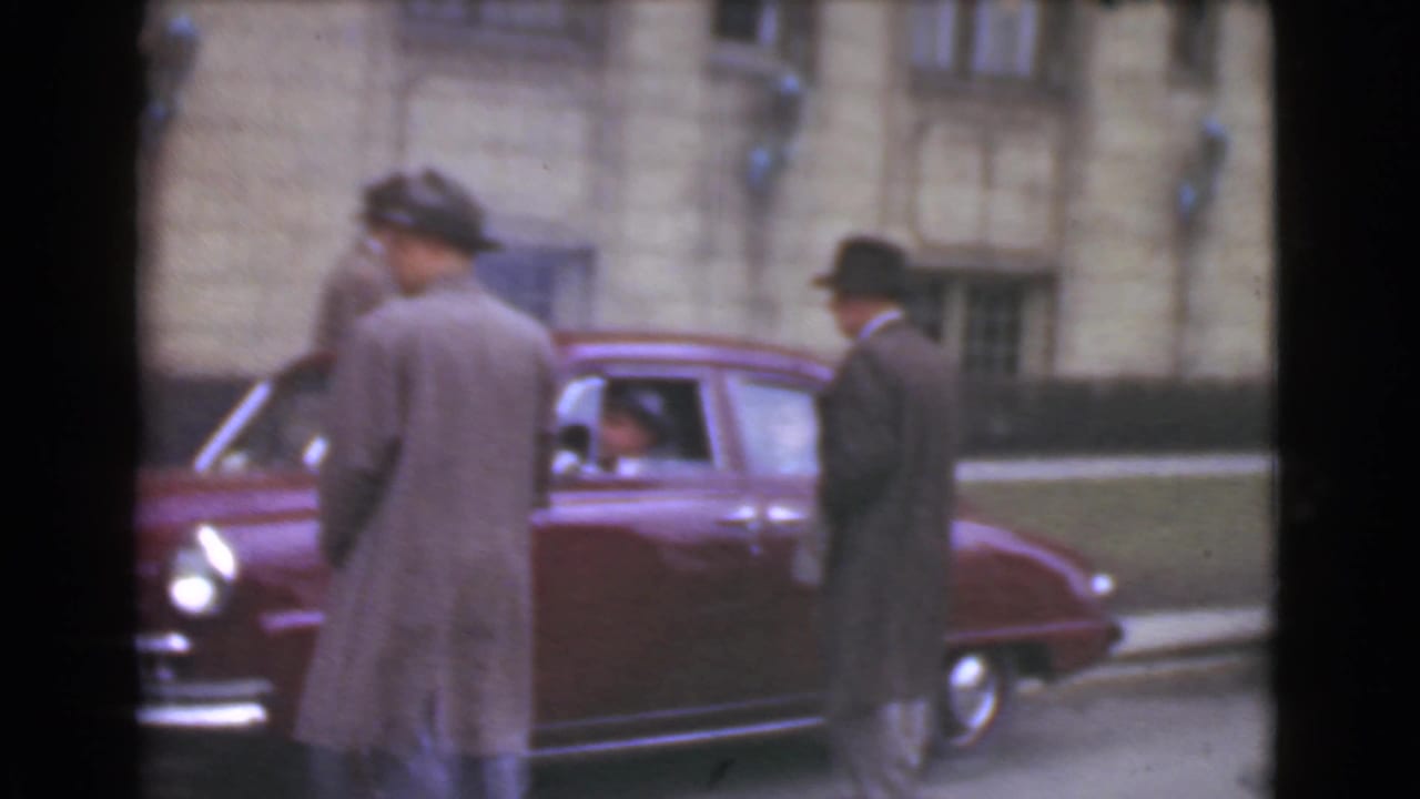 Men in overcoats and hats stand near a maroon car on a city street.