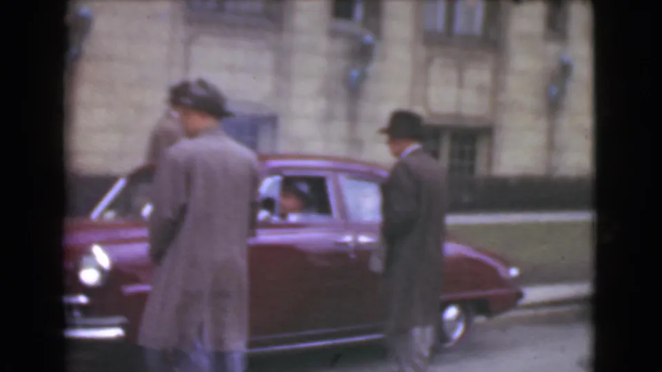In 1939 Cleveland, men in fedoras and long coats appear beside a maroon car with a stone building behind them.