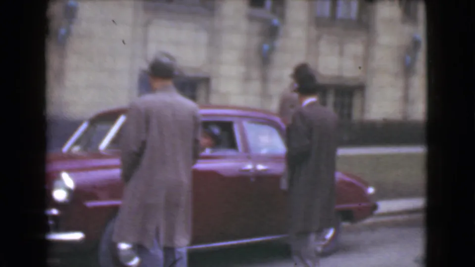 Two men in formal coats and hats stand beside a maroon car in Cleveland, with a building facade in the background.