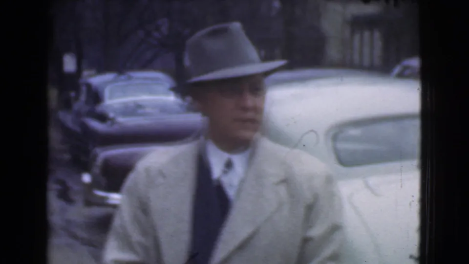 A man in a light-colored suit and fedora stands near parked cars on a Cleveland street in the 1939 home-movie footage.