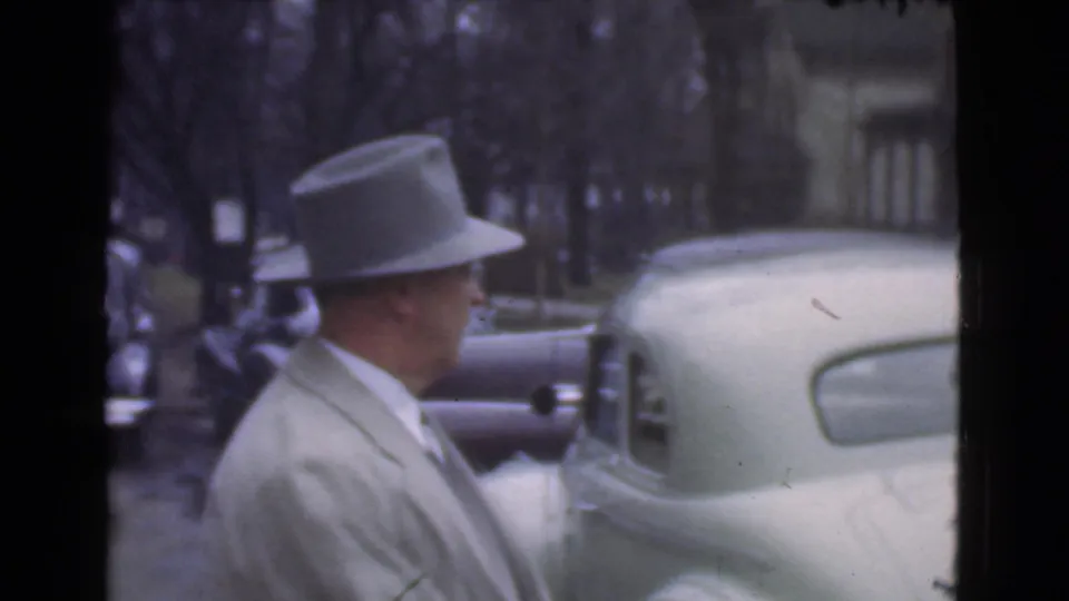 A man in a light coat and fedora turns near a light-colored parked car, with other automobiles and trees behind him in Cleveland.