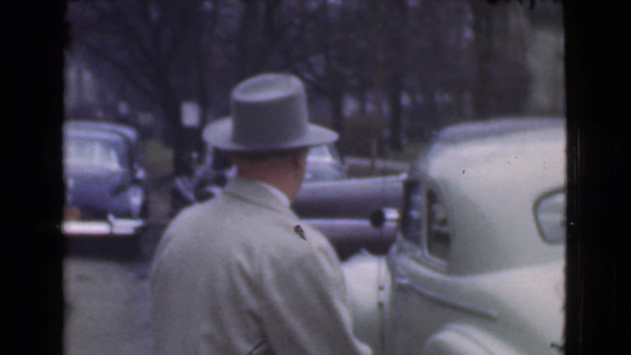 Man in a light overcoat and grey fedora seen from behind beside parked cars.