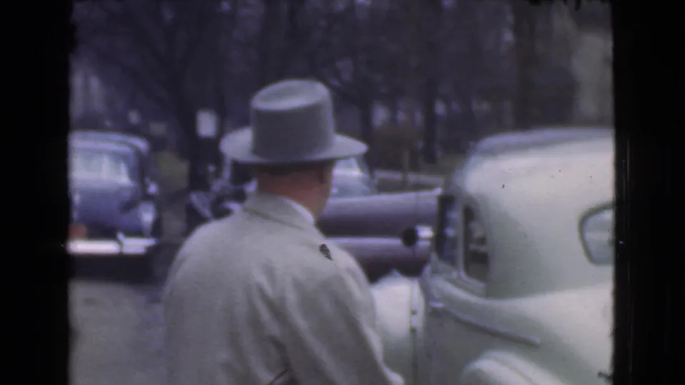 In 1939 Cleveland footage, a man in a light overcoat and grey fedora faces away near a row of parked automobiles.