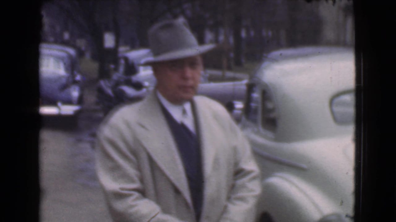 Man in a fedora and light overcoat faces the camera near a pale parked car.