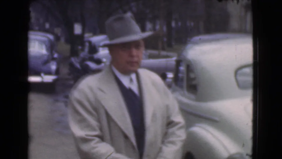 The man in a fedora, tie, and light overcoat stands near a pale rounded automobile on a Cleveland street.