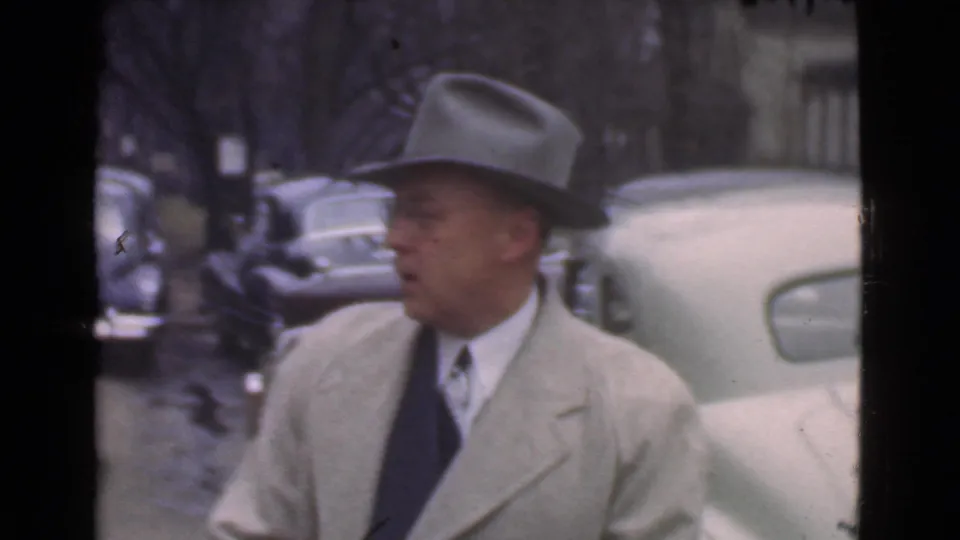 The man turns in profile near a parked car, with more rounded 1930s vehicles and trees visible behind him.