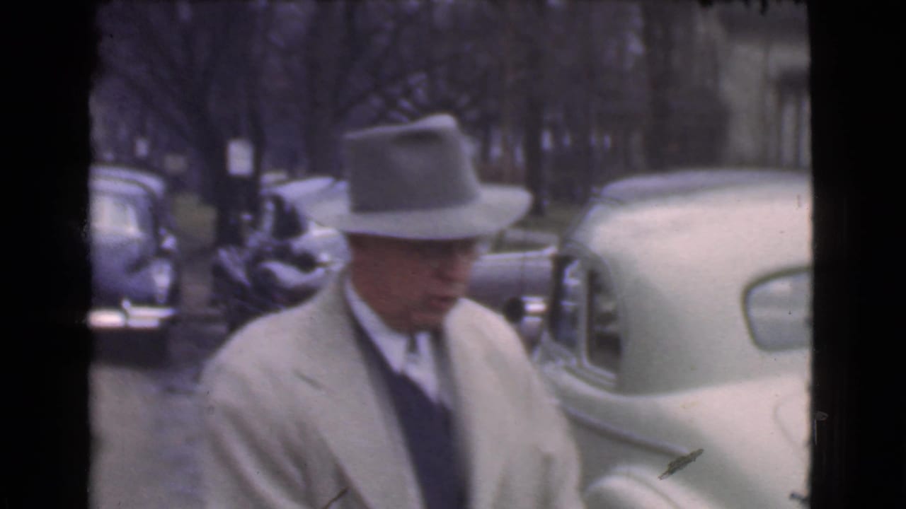 Man in a light coat and fedora walks near parked vintage cars on a city street.