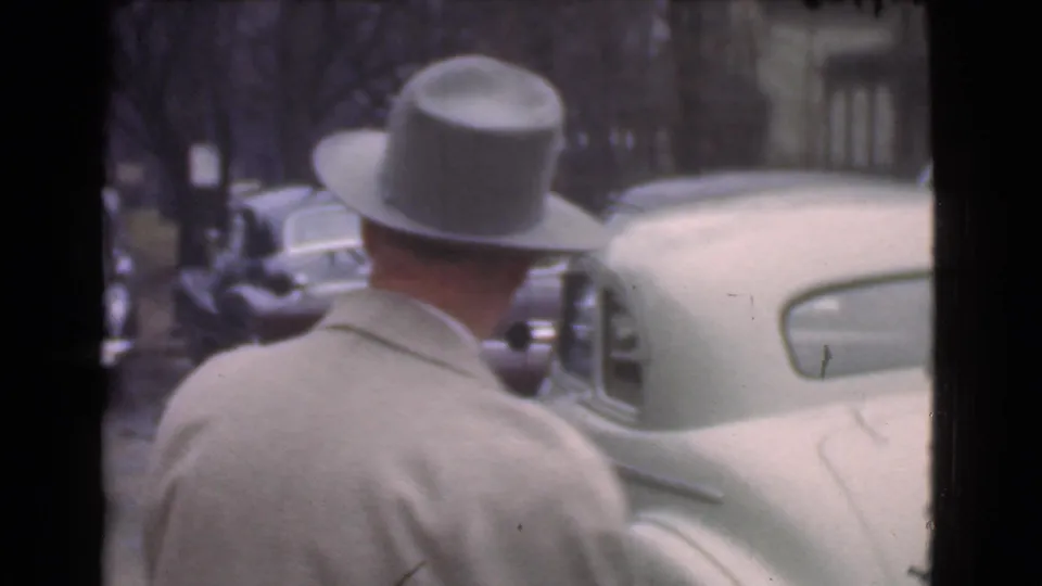 Rear view of the same man in a light coat and fedora near parked cars on a 1939 Cleveland street, with trees and buildings in the background.