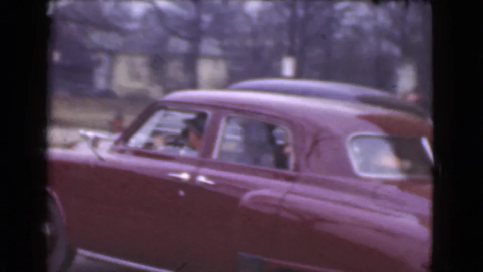 The red sedan continues past the camera in the 1939 Cleveland street footage, showing occupants through the side windows and parked cars behind.