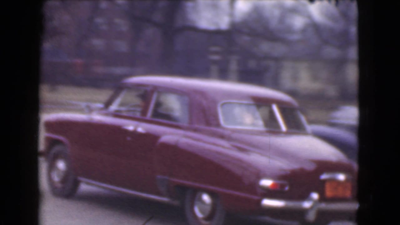 Maroon vintage sedan passing along a city street with trees and buildings behind it.