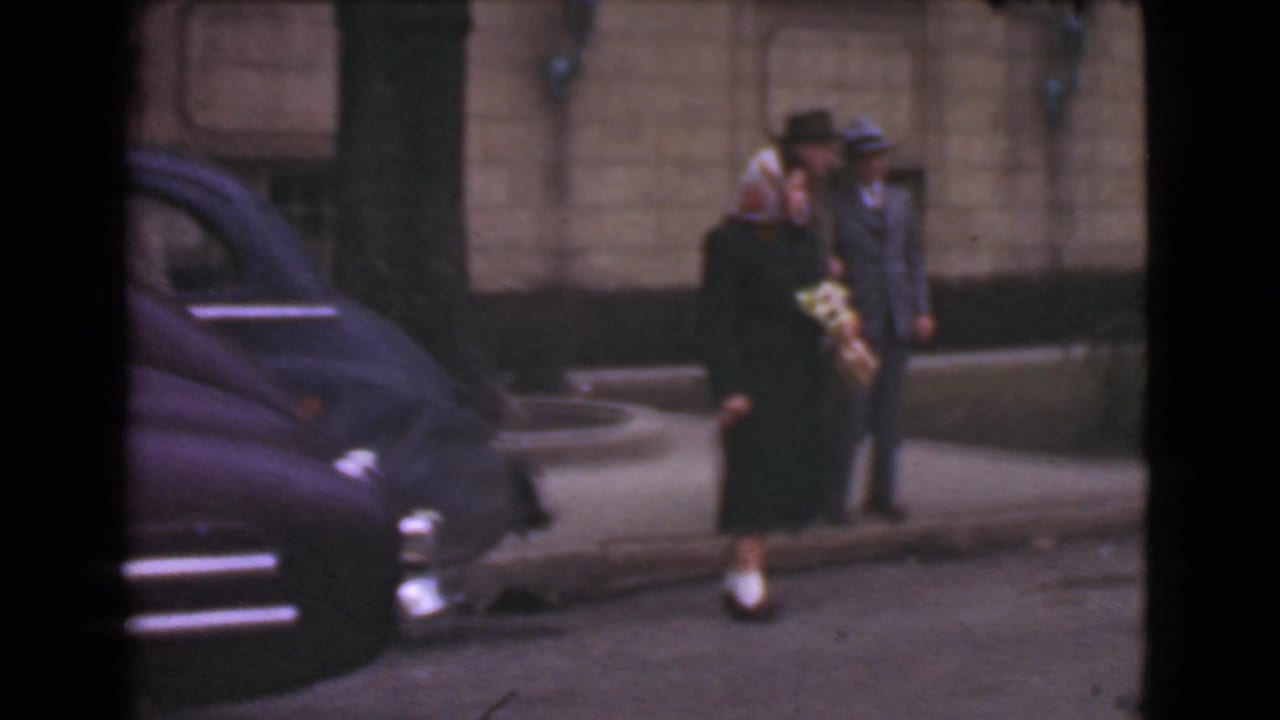 Woman holding a bouquet walks beside a suited man near a parked dark car and stone building.