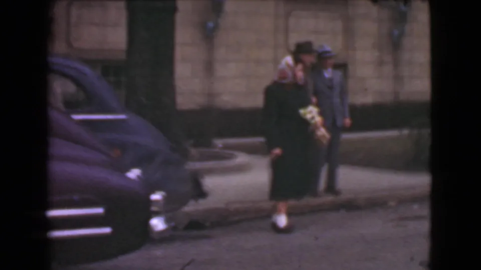 1939 Cleveland sidewalk view with a woman in a dark coat holding a bouquet beside a suited man near parked cars and a stone facade.