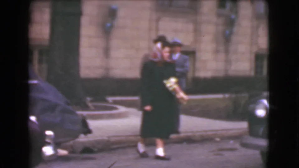 Cleveland 1939 home-movie frame showing a woman carrying a bouquet and a man in a hat walking between parked cars on a city sidewalk.