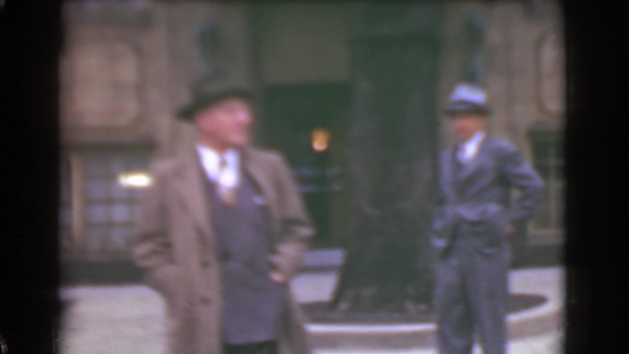Two suited men in hats stand near a city street and stone building.