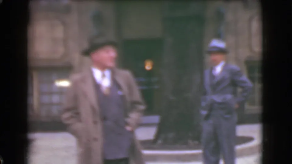 In 1939 Cleveland, two men in suits and hats wait near a sidewalk by a stone building, seen in soft 8mm color.