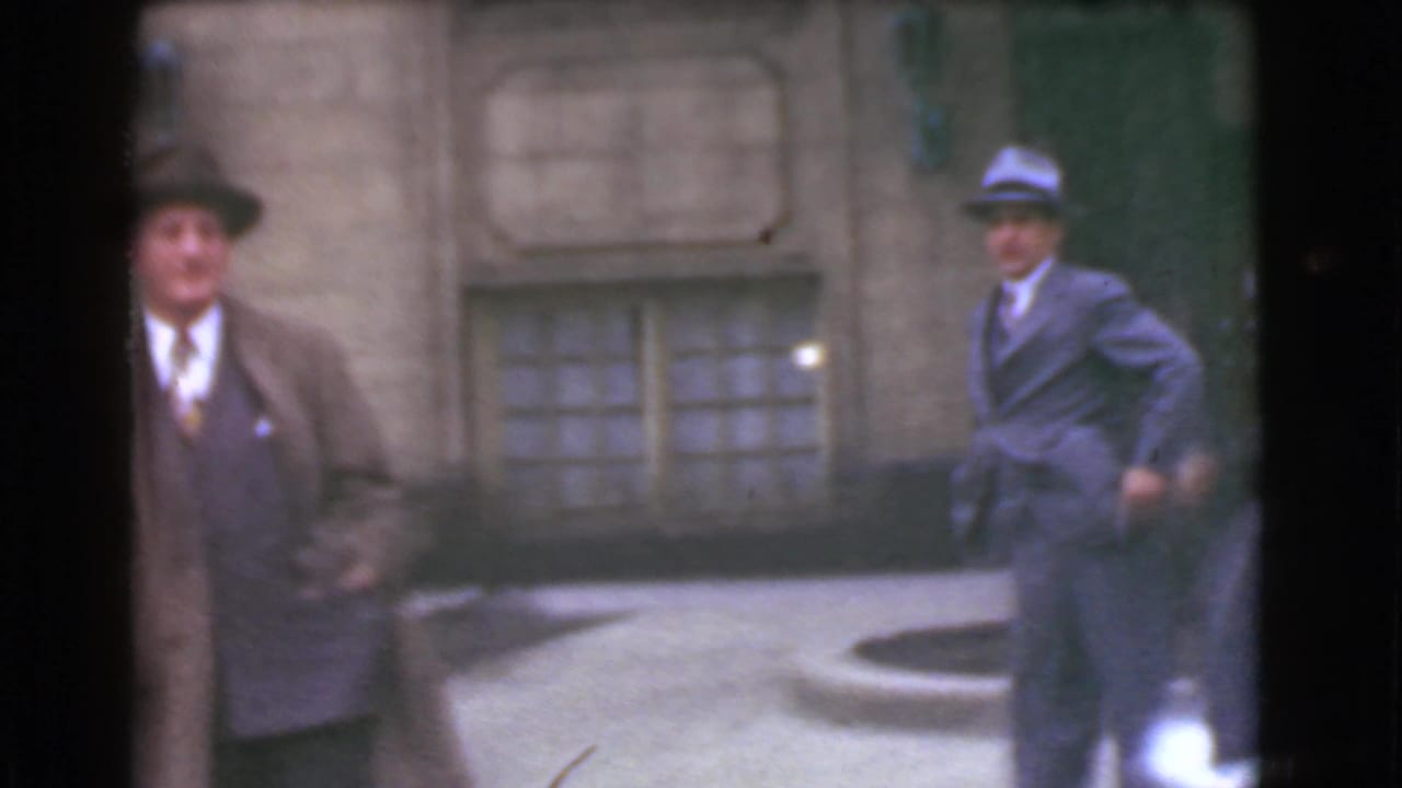 Two men in suits and hats stand apart near a stone building and street.