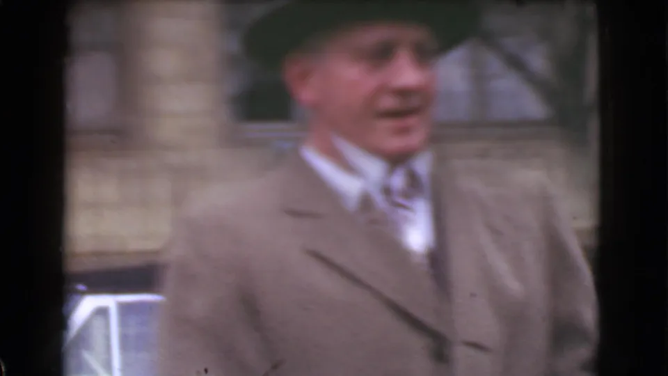 A close, soft-focus 8mm view shows a man in a brown overcoat, tie, and dark hat on a Cleveland street.