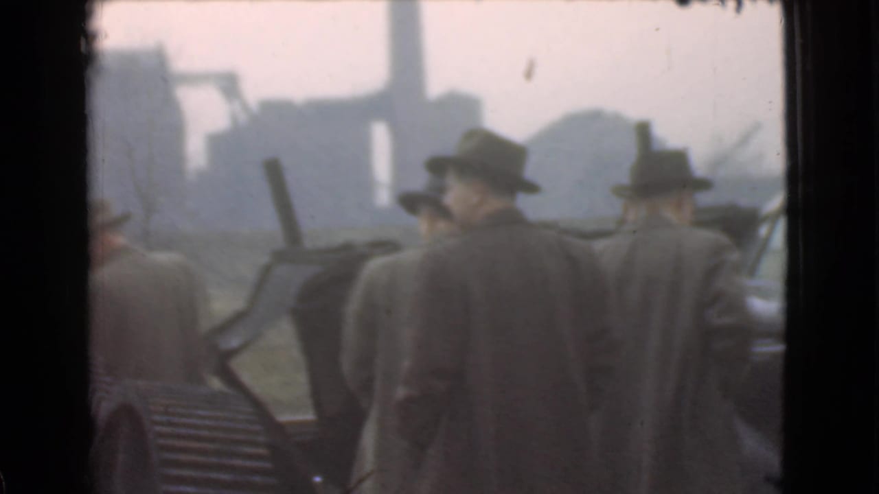 Men in hats and coats stand near a vintage car with industrial structures behind them.