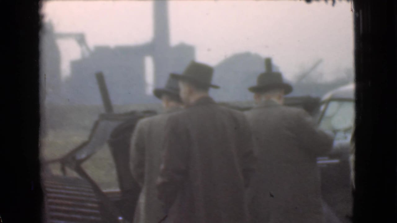 Two men in hats face away near a car, with factory-like structures in the distance.