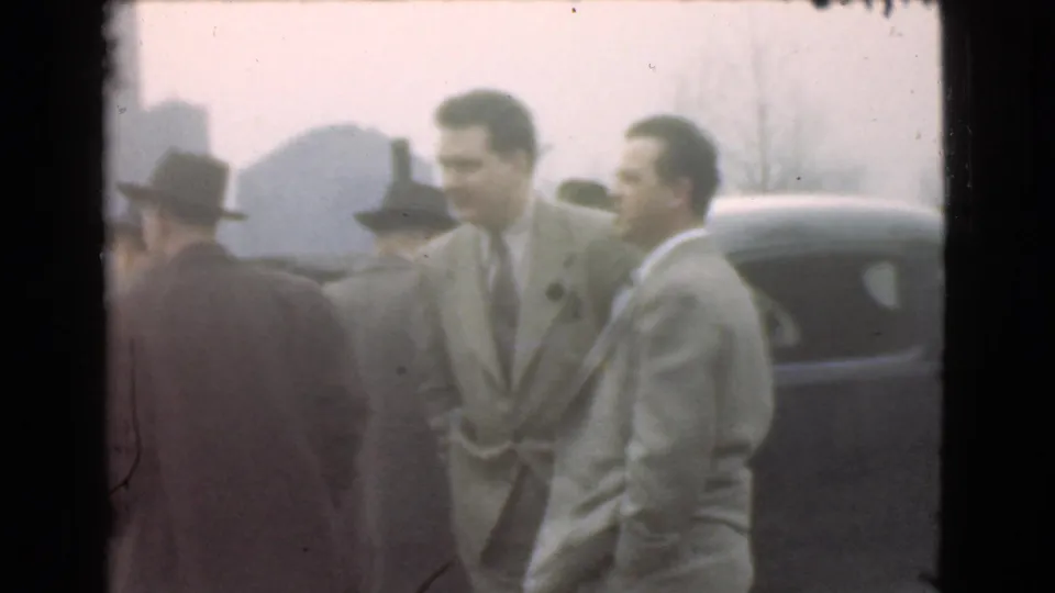 Adult men in grey suits stand near a dark rounded car in 1939 Cleveland footage, with industrial forms faintly visible behind them.
