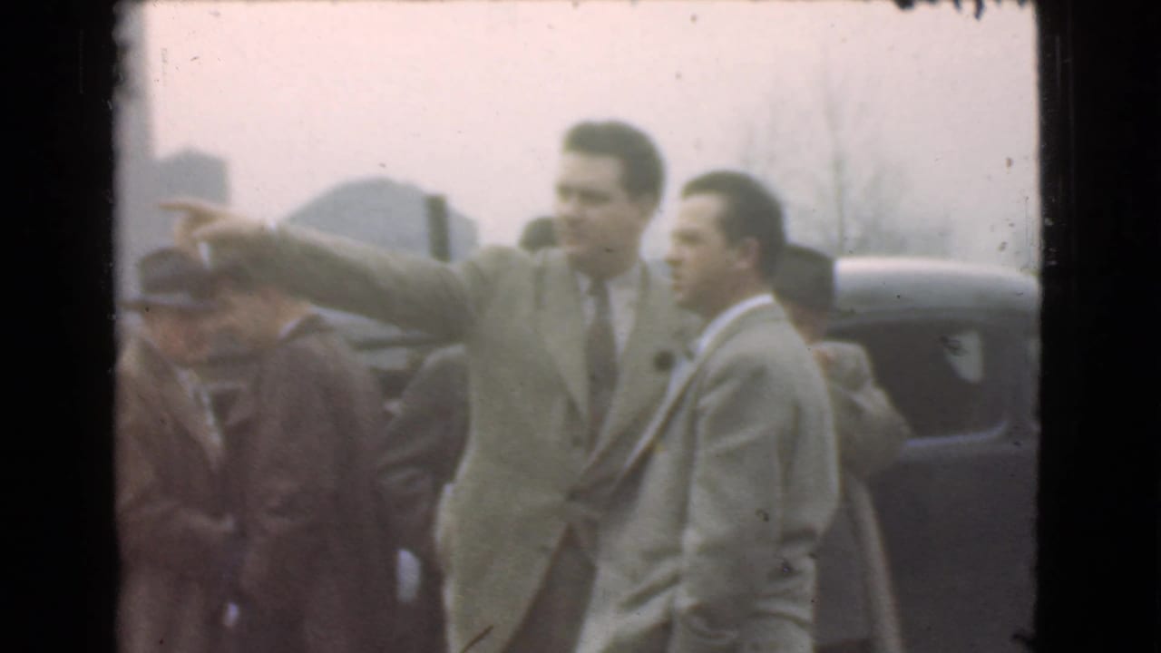 Man in a light suit points while other suited men stand near parked cars.