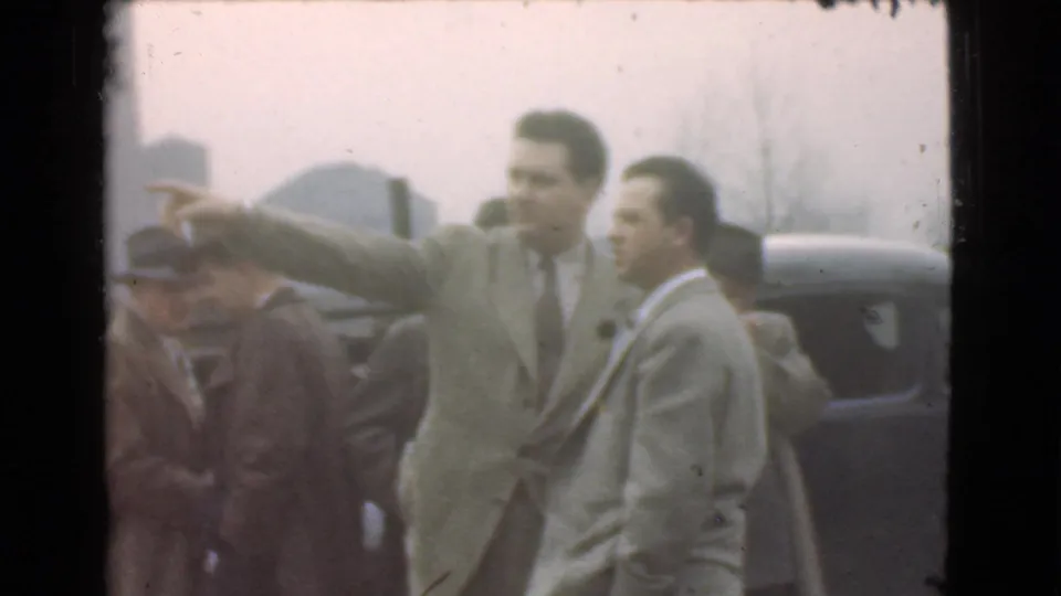 In 1939 Cleveland, a man in a light-colored suit points off frame while other adult men in suits and hats stand near parked cars.