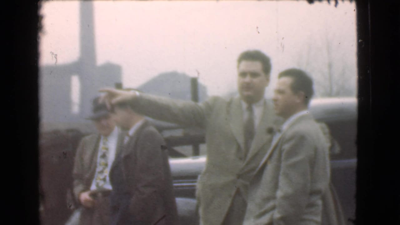 Several men stand near a car as a man in a light suit points to the left.